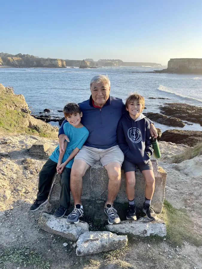 A grandfather and two boys sitting on a rock seat above the ocean in late afternoon light