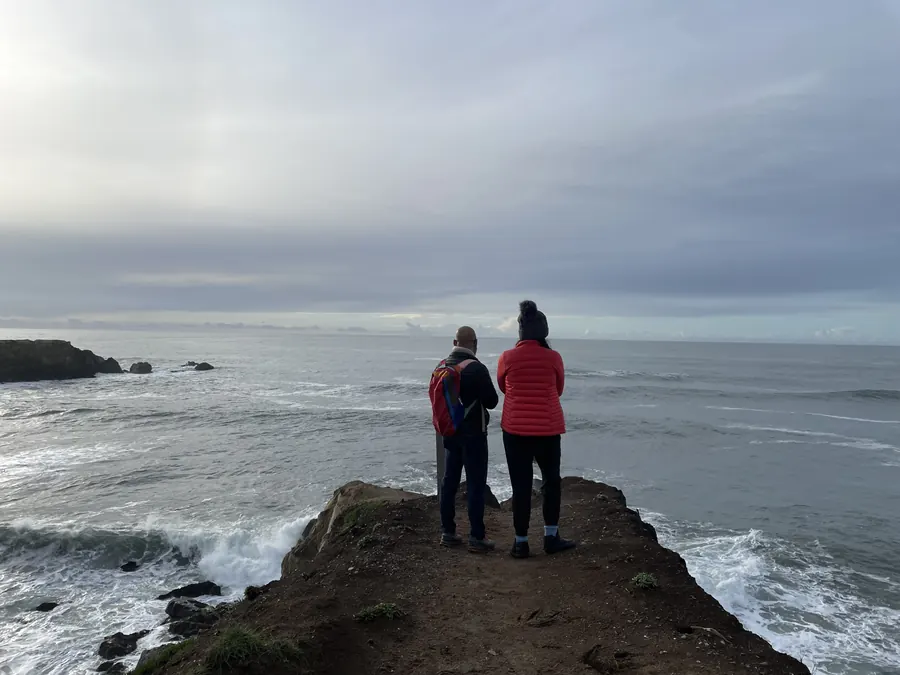 Two people standing at the end of a cliff point, looking out at a grey winter ocean