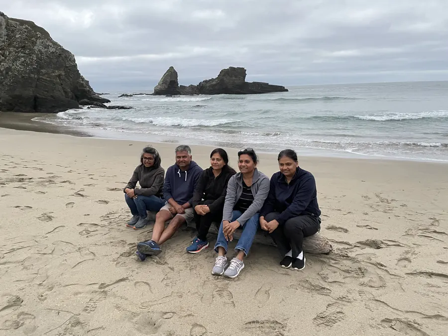 Five friends sitting together on a driftwood log at Cooks Beach, with sea stacks offshore